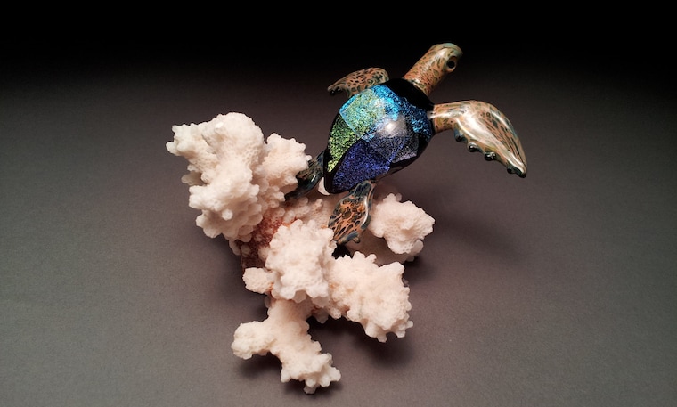 Iridescent glass sea turtle sculpture swimming above white coral